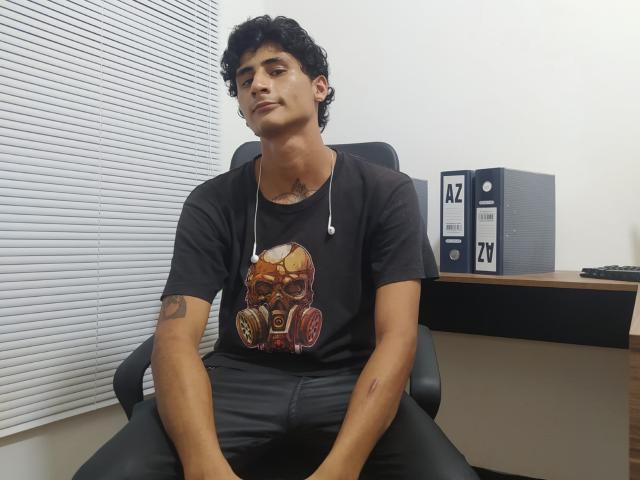CristopherHerrerX, sitting in a black chair, wearing a black t-shirt with a skull and gear graphic, black pants, and a necklace, with a tattoo on his right arm.