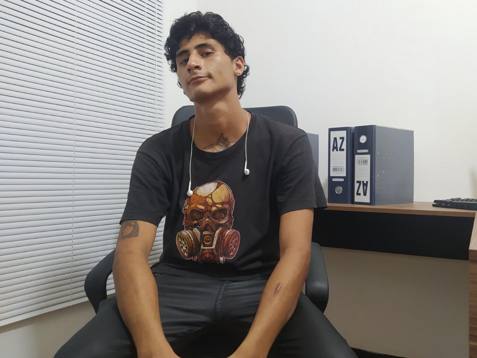 CristopherHerrerX, sitting in a black chair, wearing a black t-shirt with a skull and gear graphic, black pants, and a necklace, with a tattoo on his right arm.