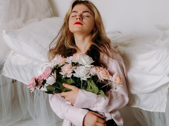 TinaRedly with eyes closed, wearing pink shirt, holding pink and white roses, sitting on white bed, red lipstick, blonde wavy hair, relaxed, soft light, gentle expression.