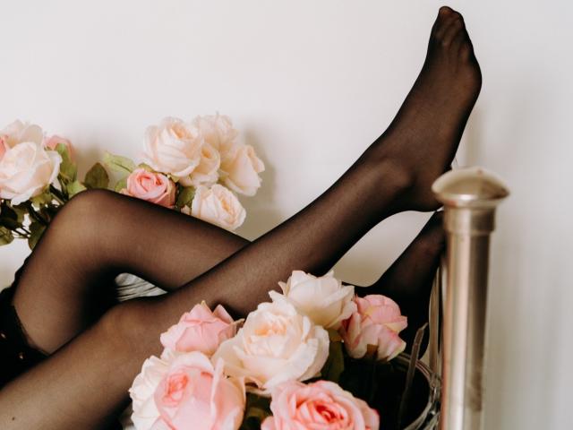 TinaRedly's legs in black sheer stockings, crossed at the ankles, resting on a bouquet of pink and white roses with a brass bedpost in the foreground.