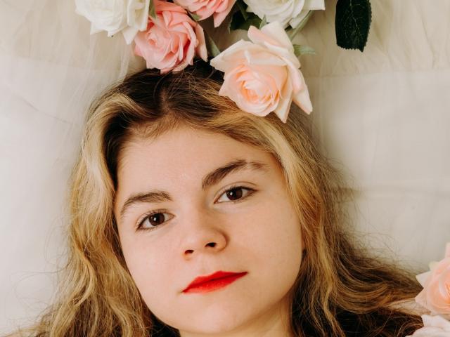 TinaRedly with blonde hair, pink roses in her hair, red lipstick, and a serious expression, lying on a white surface. Her tits are slightly visible beneath the roses.