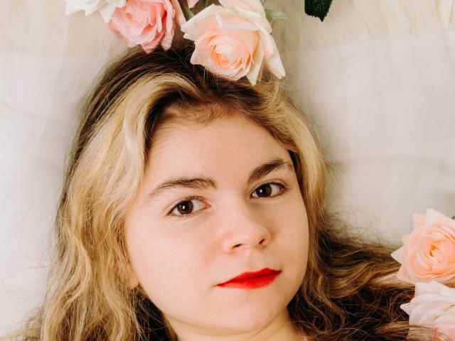 TinaRedly with blonde hair, red lipstick, and pink roses in her hair, lying down, looking at the camera, showing her face and upper chest.