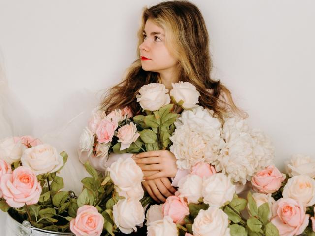TinaRedly with wavy blonde hair, red lipstick, and white dress, holding pink and white roses, looking to the side, surrounded by flowers. Soft, romantic vibe.