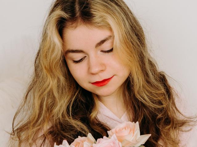 TinaRedly with wavy blonde hair, closed eyes, red lipstick, holding pink roses, wearing a pink top, soft smile, white background. Her tits are not visible.