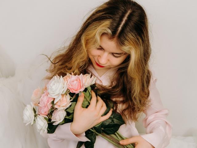 TinaRedly with wavy blonde and brown hair, wearing a pink shirt, holding a bouquet of pink and white roses, looking down with a gentle smile.