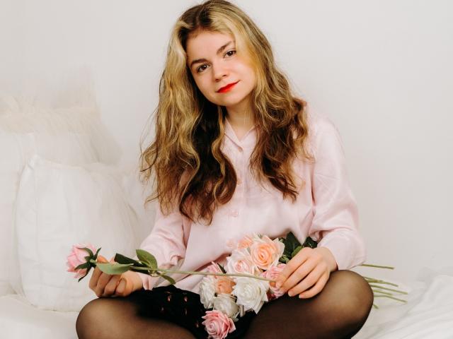 TinaRedly with wavy blonde hair, pink shirt, black tights, holding pink roses, sitting on white bed, smiling with red lipstick, hands on roses, legs crossed.