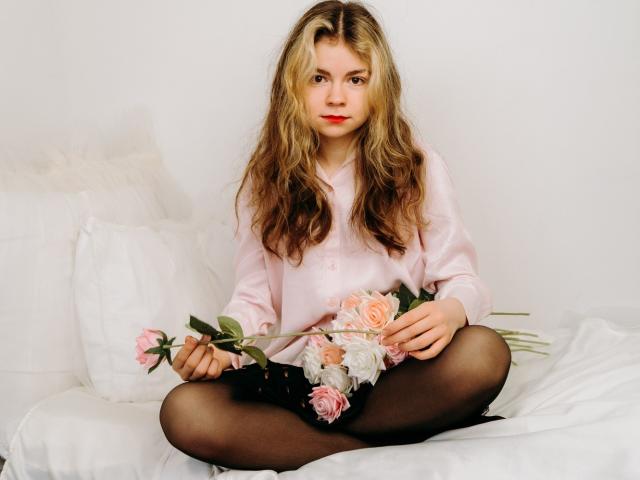 TinaRedly, com cabelos loiros ondulados, camisa rosa, collant preto, sentada de pernas cruzadas em uma cama branca, segurando rosas rosa e brancas, com uma expressão séria e um leve batom vermelho..