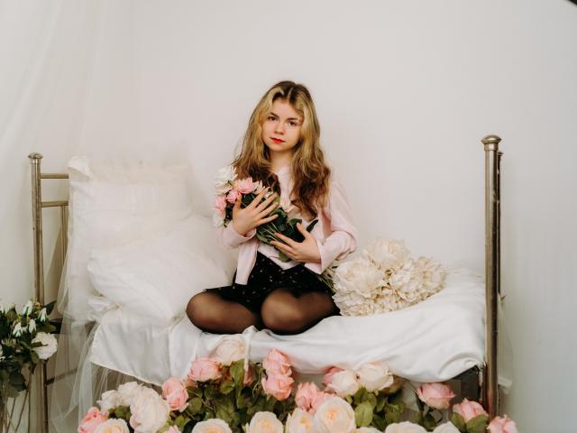 TinaRedly with long blonde hair, pink shirt, black polka dot skirt, holding pink roses, sitting on white bed, surrounded by pink roses, black tights, white pillows, brass bed.