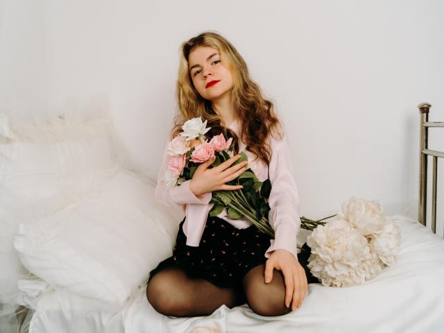 TinaRedly with long blonde hair, pink shirt, black polka-dot skirt, and black stockings, holding pink roses, sitting on white bed with white pillows and large white peonies.
