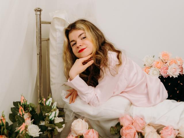 TinaRedly with wavy blonde hair, red lipstick, and pink shirt, lying on bed, head resting on hand, surrounded by pink and white roses, smiling softly.