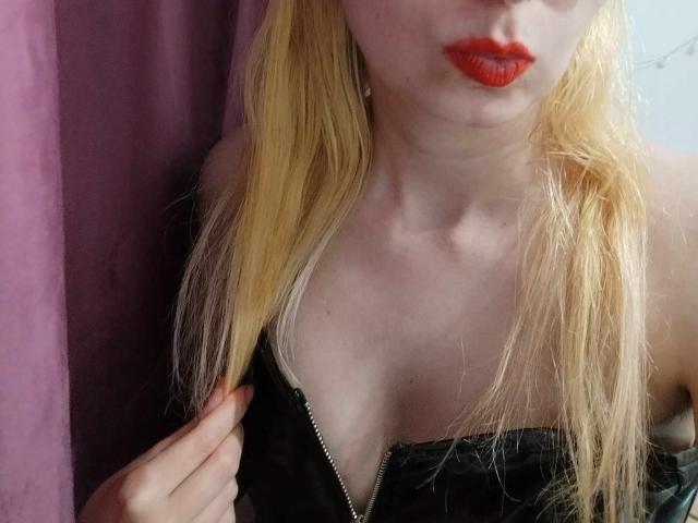 AlexandraSweet mit blonden Haaren, rotem Lippenstift, einem schwarzen Kleid mit Reißverschluss, die ihre Haare hält, ihre Brüste zeigt, blasser Haut und einem verführerischen Blick..