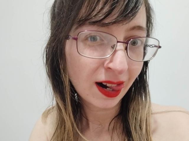AlexandraSweet with messy black-brown hair, red lipstick, and glasses, has blue eyes and a slightly open mouth showing a small bit of her teeth. She's wearing a strapless top.