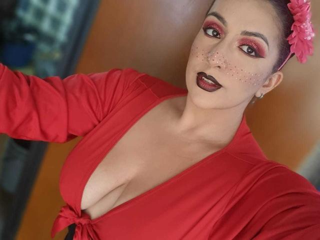 AndreaSharick has pink flower in hair, red makeup, red dress, deep cleavage, black lipstick, freckles, red eyes, holding phone, orange background, medium shot, showing tits.