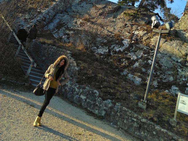RachelHotTs has long brown hair, a tan coat, black pants, and yellow boots, standing on a gravel path with rocky hills and a metal signpost in the background.