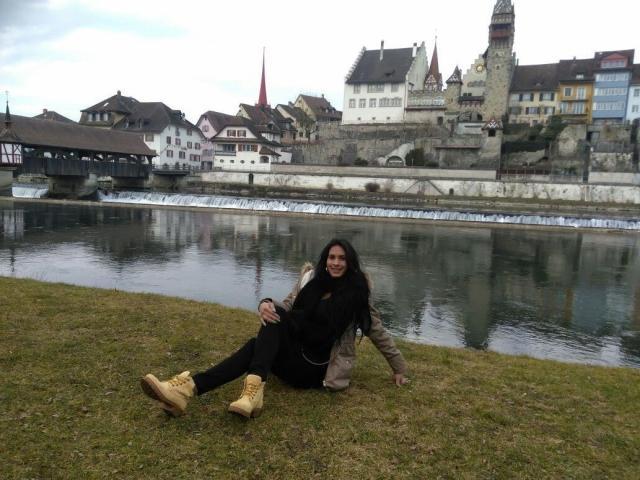 RachelHotTs sits on grass by a river, wearing a black top, black pants, tan boots, and a beige jacket. Her ass is slightly visible, and she has long dark hair.