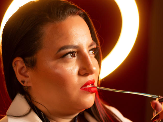 SkayLaraWalker mit rotem Lippenstift, die einen Make-up-Pinsel vor ihren leicht geöffneten Mund hält, dunkles Haar, braune Augen, weißer Kragen, warmer Lichtkreis im Hintergrund..