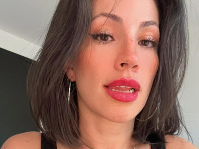 Liliysweet has medium brown, slightly wavy hair, red lipstick, and wears hoop earrings, with visible teeth and subtle eye makeup, looking slightly to the side.