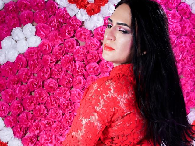 IrisDurand with long black hair, red lace shirt, and pink eyeshadow, standing against a wall of pink and white roses, looking back with a slight smile.