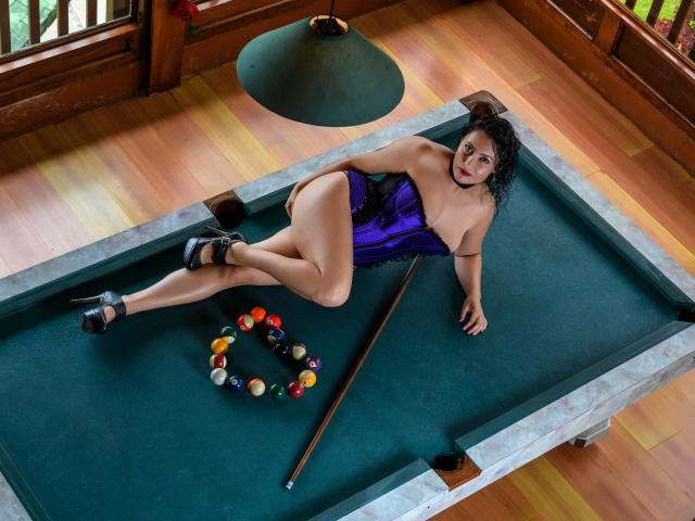 YennyFort with curly black hair, wearing a purple and black corset, black heels, lying on a pool table, legs crossed, pool balls in a triangle, cue stick.