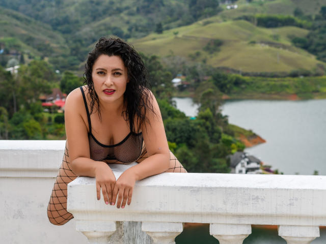 YennyFort leans on a white balcony, wearing a sheer black top showing her big boobs, fishnet stockings, and wet curly hair, with a sexy smile.