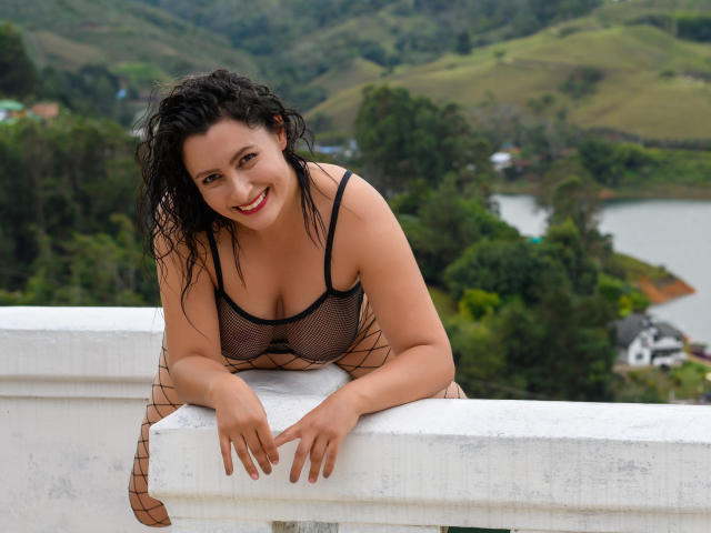 YennyFort with curly black hair, smiling, wearing a black mesh dress, leaning on a white railing, showing her boobs, with a green, hilly landscape in the background.