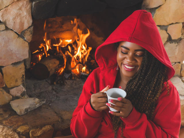 DanielaBrown with dark skin, curly brown hair, red hoodie, smiling, holding white cup, sitting by stone fireplace with bright orange flames. Her tits are visible under the hoodie.