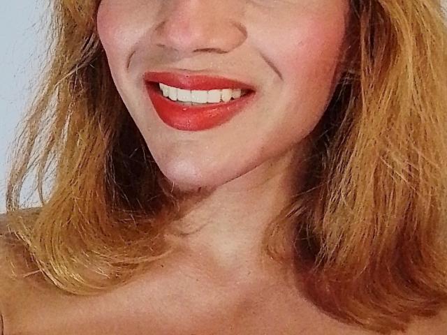 TatyLorenX a du rouge à lèvres, elle sourit en montrant ses dents blanches, et a des cheveux ondulés châtain clair. On voit son cou et le haut de sa poitrine, elle ne porte pas de vêtements. Sa peau est d'un rose pâle, et on aperçoit légèrement sa mâchoire..