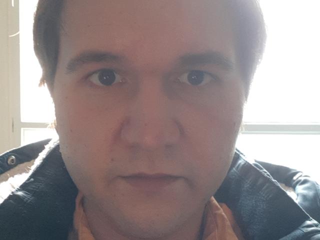 NectarineFad has a chubby face with light skin, brown hair, and a slightly open mouth. He's wearing a dark jacket over an orange shirt. Sunlight is in the background.