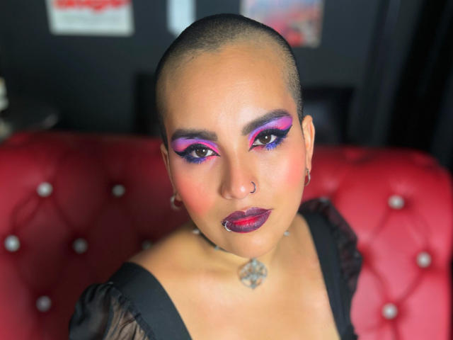 SilenaLee with shaved head, purple and pink eye makeup, red lips, nose ring, black dress, and necklace, sitting on red tufted couch. Boobs partially visible, ass not visible.
