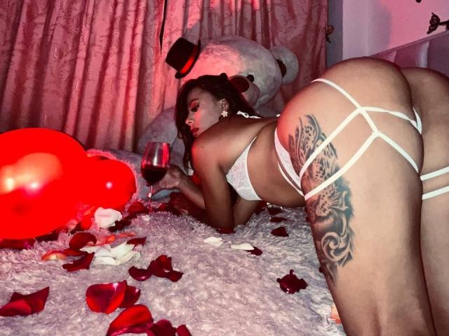 Amberv, con lencería blanca, sosteniendo una copa de vino tinto, a cuatro patas, mostrando su trasero y espalda tatuados, con pétalos de rosa y globos rojos sobre una cama peluda..