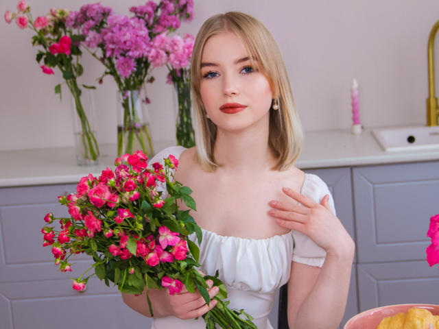 NicoleBrian with blonde hair, blue eyes, and red lipstick, wearing a white off-shoulder dress, holding a pink rose bouquet, touching her chest, in a kitchen with flowers.