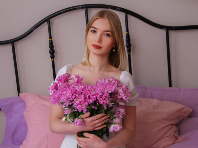 NicoleBrian met blond haar, roze bloemen, een witte jurk, die een boeket vasthoudt, roze en paarse kussens, een zwart metalen hoofdeinde, schattig, aantrekkelijk, kleine borsten, roze lippen, een lichte glimlach, zacht licht..