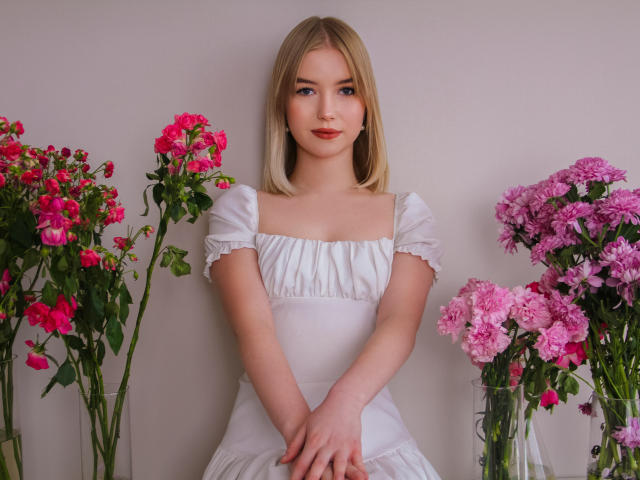 NicoleBrian with blonde hair, wearing a white dress, standing between two clear glass vases with pink and purple flowers, hands gently clasped in front.