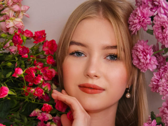 NicoleBrian's face with blonde hair, blue eyes, and orange lipstick, surrounded by pink and red flowers, resting her chin on her hand. She's wearing a pearl earring.