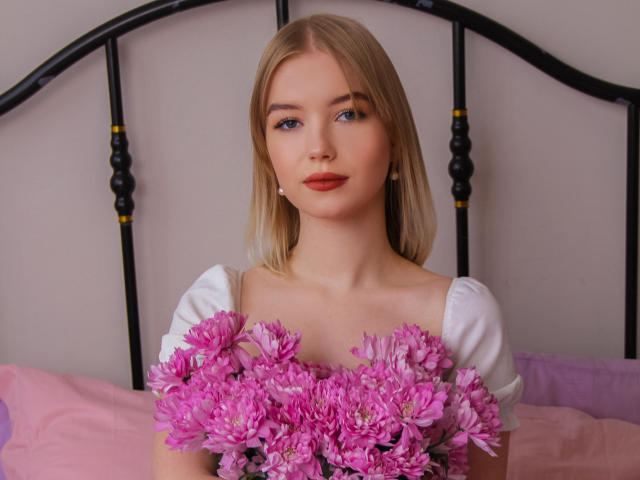 NicoleBrian has blonde hair, blue eyes, and red lipstick. She's holding pink flowers in front of her white dress, standing in front of a black metal headboard.