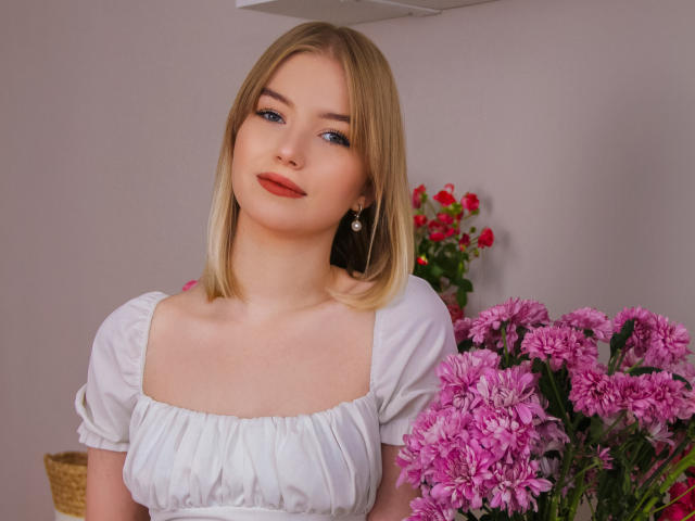 NicoleBrian with blonde hair, blue eyes, red lipstick, wearing a white dress, holding pink chrysanthemums, small earrings, subtle smile, soft light, warm indoor setting.