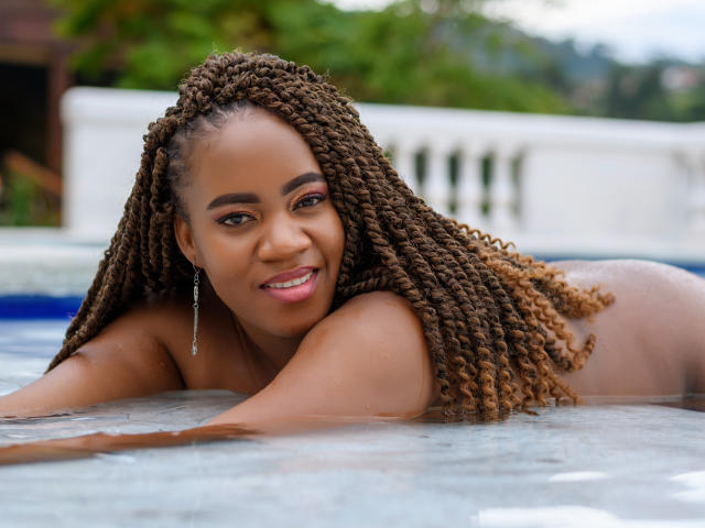 DanielaBrown with brown braided hair, smiling, lying in a pool, topless, showing her ass, with water up to her shoulders, wearing a silver earring, looking at the camera.