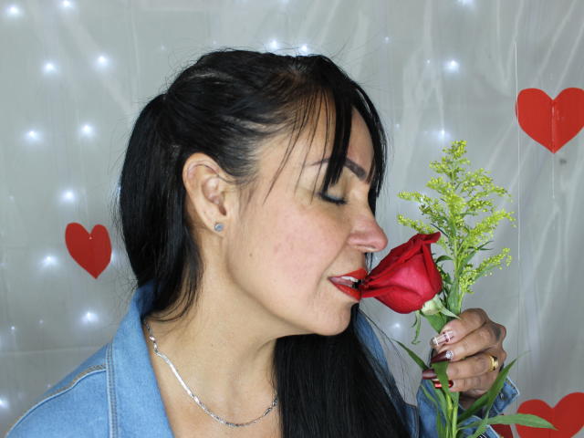 Laila Castillo, com cabelo preto, batom vermelho, piercing no nariz e calça jeans azul, cheirando uma rosa vermelha com caule verde, usando um colar de prata e brincos azuis..