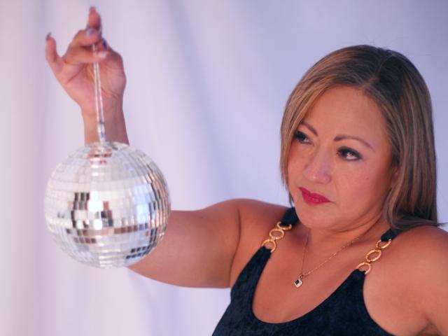 ChristinMoore with blonde hair, red lipstick, holding a shiny mirror ball, wearing a black tank top with gold straps, looking slightly to the side.