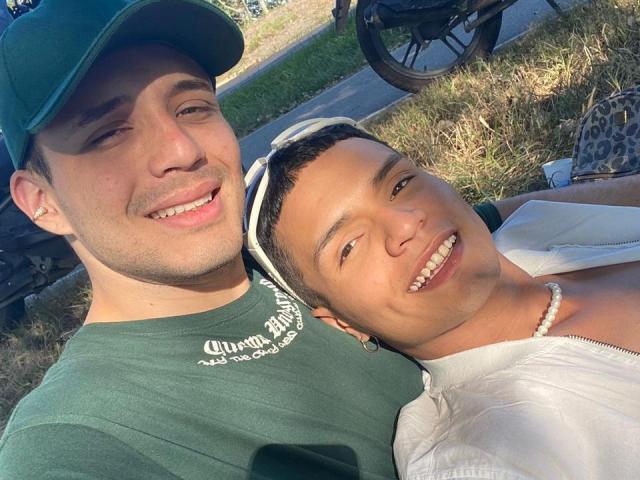 DuoLatinHoty with short brown hair and white shirt, smiling, leaning on DuoLatinHoty with green cap and green shirt, both with cute smiles, relaxed in sunlight.