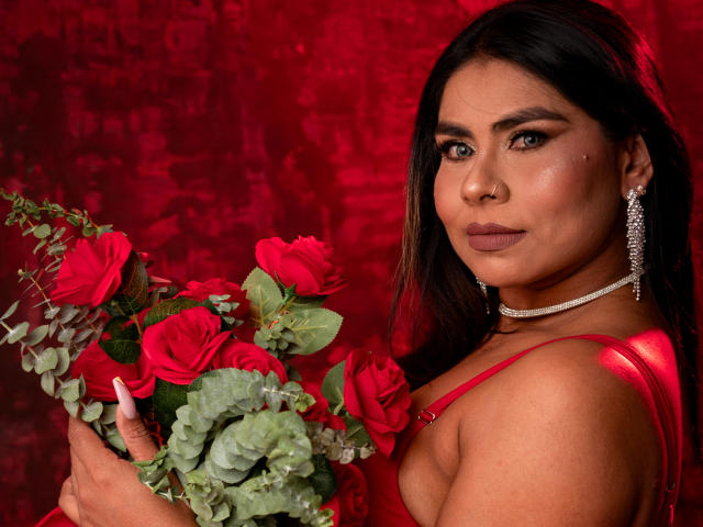 SusanClaire con rose rosse, che tiene un bouquet, indossa un abito rosso lucido, una collana e orecchini di diamanti, guarda la fotocamera, ha i capelli castani, la pelle media, trucco e un piercing al naso, su sfondo rosso..