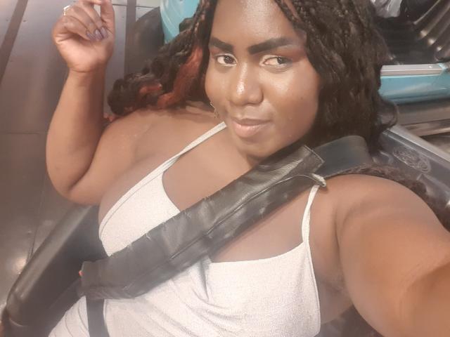 Dilay with dark skin, braided hair, and red highlights, wearing a white tank top, showing her boobs, seated with a black strap across her chest, smiling.