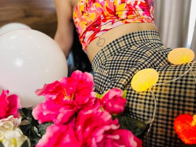 As nádegas de LisaAlisia, vestindo uma saia quadriculada preta e amarela com botões amarelos, e uma blusa vermelha com estampas florais, têm uma tatuagem de coração na parte inferior das costas, cercada por flores rosa..