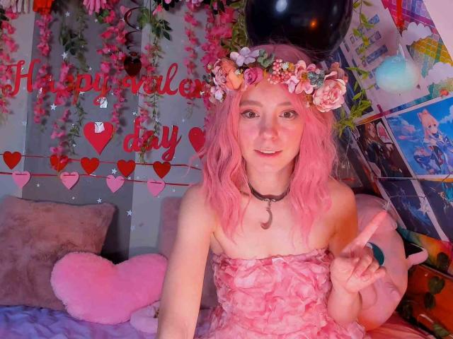 MissAstravert with pink hair, flower crown, strapless pink dress, black choker, and double peace sign, sitting on bed with pink heart pillow, smiling. Boobs not visible.