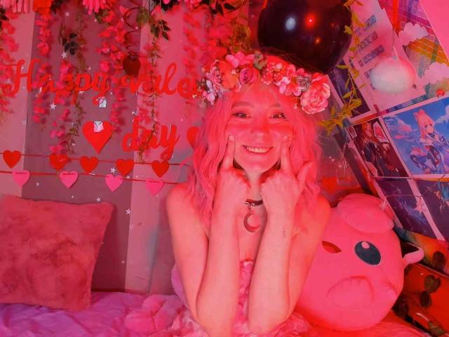 MissAstravert mit rosa Haaren, Blumenkranz, lächelnd, Hände auf den Wangen, rosa Kleid, rosa Zimmer, rosa Kirby-Plüschtier, rote Herzdekorationen, rosa Beleuchtung, süß, sexy, flauschiges rosa Kissen..