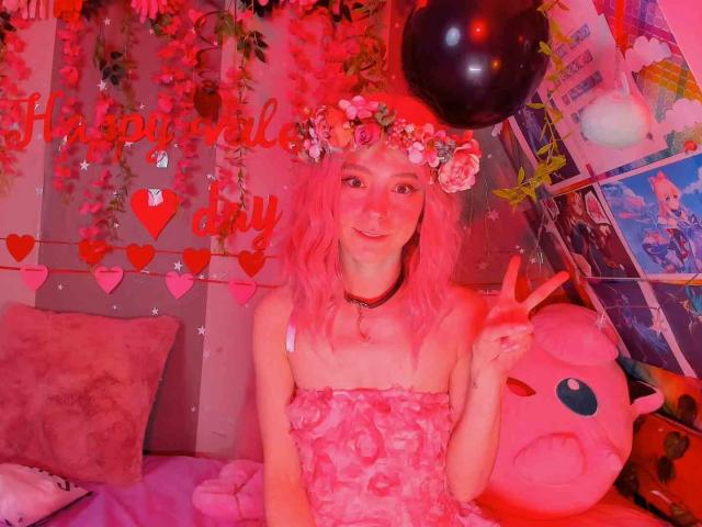 MissAstravert with pink hair, flower crown, pink strapless dress, making a peace sign, next to a pink Kirby plushie, in a pink-lit room with a black balloon.