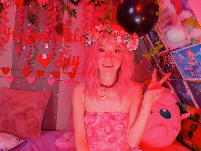 MissAstravert with pink hair, flower crown, crescent moon necklace, pink strapless dress, making a peace sign, in a pink-lit room with a black balloon and pink plushie.