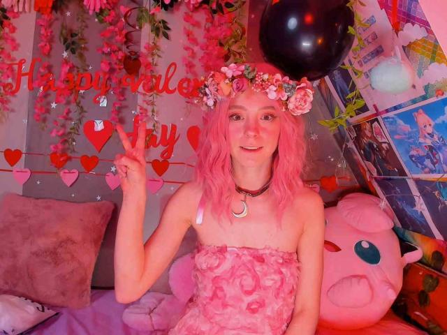 Pink-haired MissAstravert with flower crown, wearing a pink, ruffled, strapless top, showing a peace sign, her small tits visible, surrounded by pink balloons and plush toys.