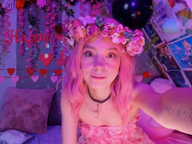 Pink-haired MissAstravert with flower crown, pink dress, and black choker, smiling at camera, showing small tits, cute face, and tattoo on arm, in pink-lit room.