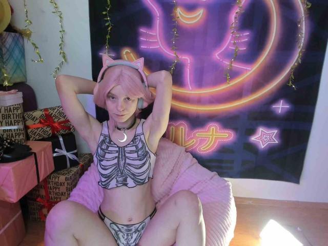 MissAstravert with pink hair, cat ears, and a skeleton crop top, sitting on a pink pillow, legs spread, showing her small tits and pussy, arms raised, neon sign behind.